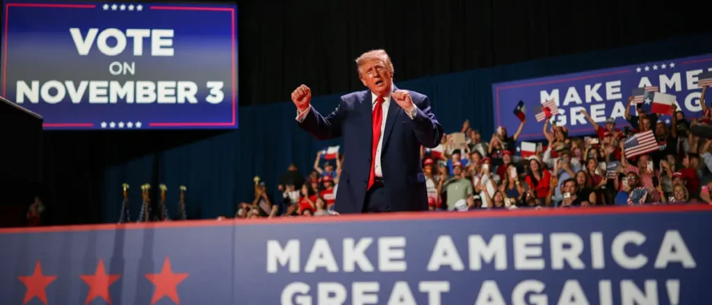 The image shows a political rally with a prominent sign encouraging people to vote on November 3 and another sign reading "MAKE AMERICA GREAT AGAIN!" with a crowd of supporters in the background.