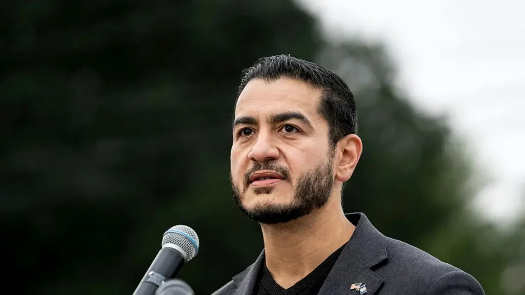 The image shows a man speaking into a microphone, dressed in a suit, with a serious expression, set against a backdrop of trees.