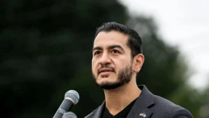 The image shows a man speaking into a microphone, dressed in a suit, with a serious expression, set against a backdrop of trees.