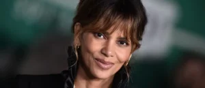 The image shows a person with a warm smile, wearing a black outfit and gold earrings, set against a blurred background.