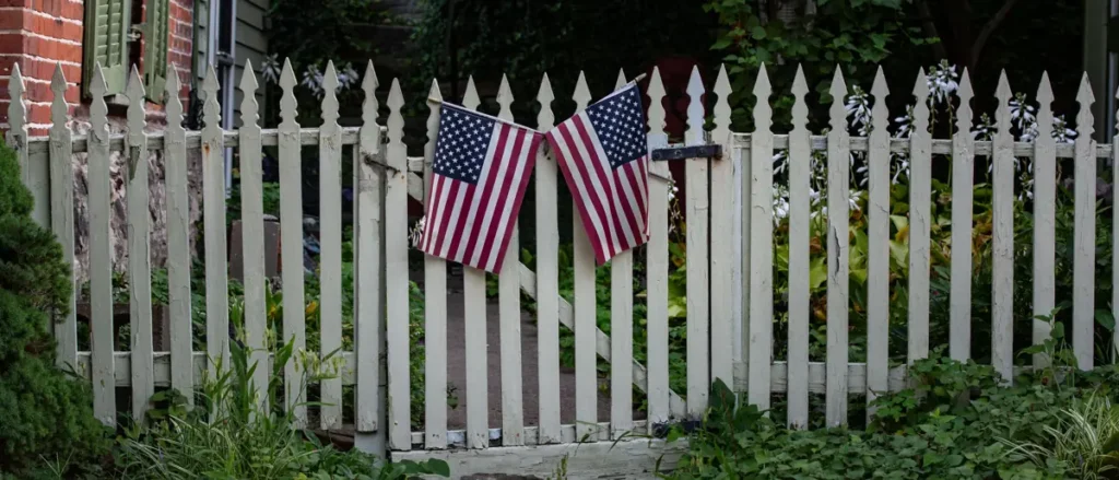 A white picket fence with two American flags is seen in front of a garden with lush greenery.