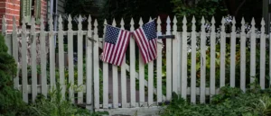 A white picket fence with two American flags is seen in front of a garden with lush greenery.