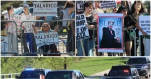Protesters gather behind a barricade, holding signs and a poster, as vehicles drive by on a road.