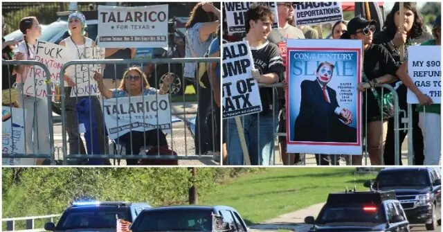 Protesters gather behind a barricade, holding signs and a poster, as vehicles drive by on a road.