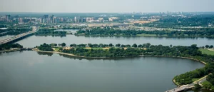 The image shows an aerial view of a cityscape with a large body of water in the foreground, surrounded by greenery and urban infrastructure.