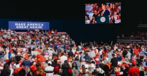 The image depicts a large rally with a crowd gathered, many wearing red hats, and a stage featuring a speaker and a large screen displaying the same scene.