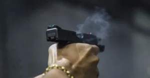A close-up of a hand holding a handgun, with visible smoke and a gold chain bracelet.