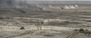 The image depicts a desert battlefield with military vehicles and smoke rising from explosions in the distance.