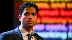 A man in a suit and tie stands at a microphone, appearing serious and focused, with a colorful, blurred background.