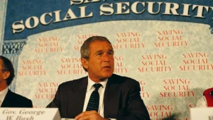 The image shows a man in a suit speaking at a podium with a backdrop that reads "SAVING SOCIAL SECURITY."