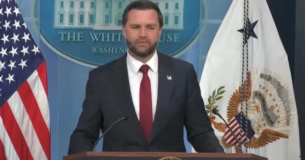 A man in a suit and tie stands at a podium in front of the White House seal and American flags.