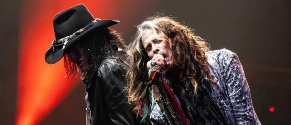 Two musicians perform on stage, one wearing a black hat and the other holding a microphone, with vibrant stage lighting in the background.