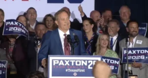A politician stands at a podium with supporters behind him, holding signs that read "PAXTON 2026" and "TexasFirst."
