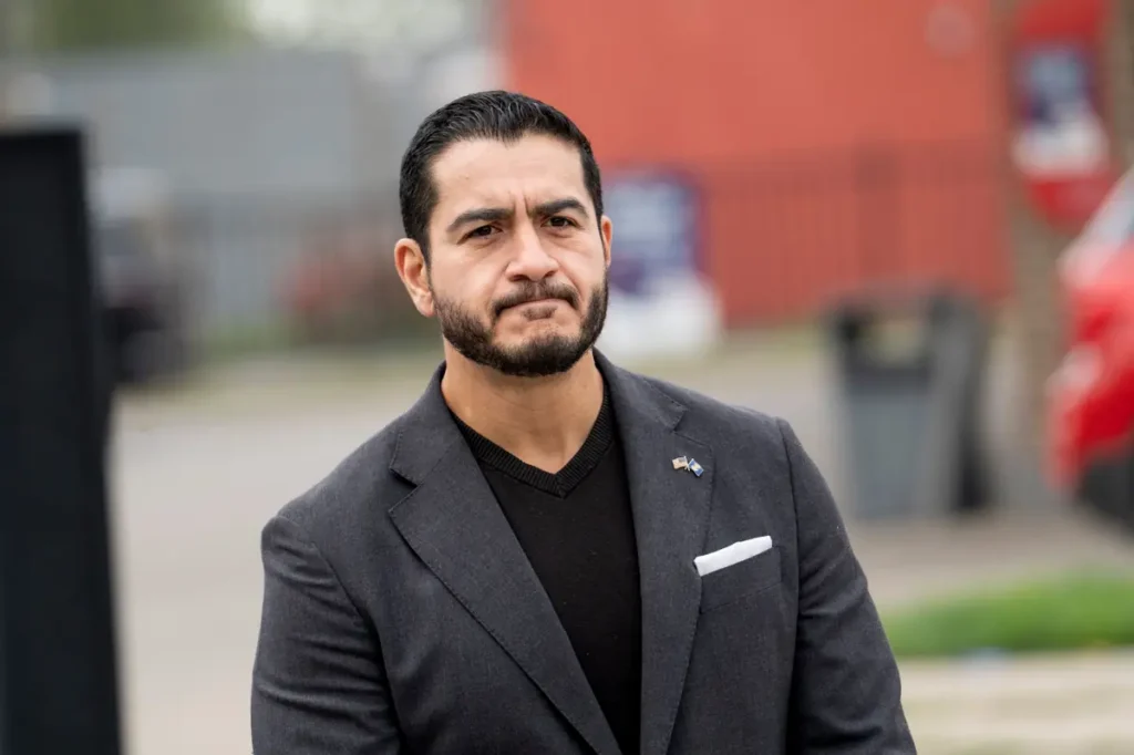 The image shows a man in a suit standing outdoors, with a serious expression and a flag pin on his lapel.