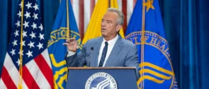 A man in a suit stands at a podium with the Department of Health and Human Services seal, delivering a speech in front of American and Department flags.