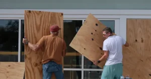 Two individuals are working together to secure large plywood sheets to a building's windows, likely as a precautionary measure.