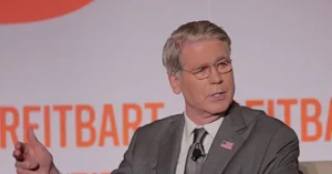 The image shows a man in a suit and tie speaking at a podium with the word "REITBART" visible in the background.