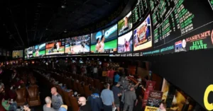 The image shows a bustling sportsbook with numerous screens displaying live sports action and a crowd of people engaging in various activities.