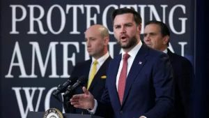 A man in a suit stands at a podium with microphones, speaking in front of a backdrop that reads "PROTECTING AMERICA'S WORKERS."