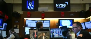 The image shows a trading floor at the New York Stock Exchange (NYSE) with traders working at their stations, some wearing red hats, and monitors displaying financial data.