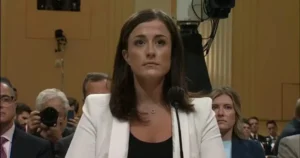 A woman in a white blazer speaks at a formal event, with a microphone in front of her and an audience in the background.