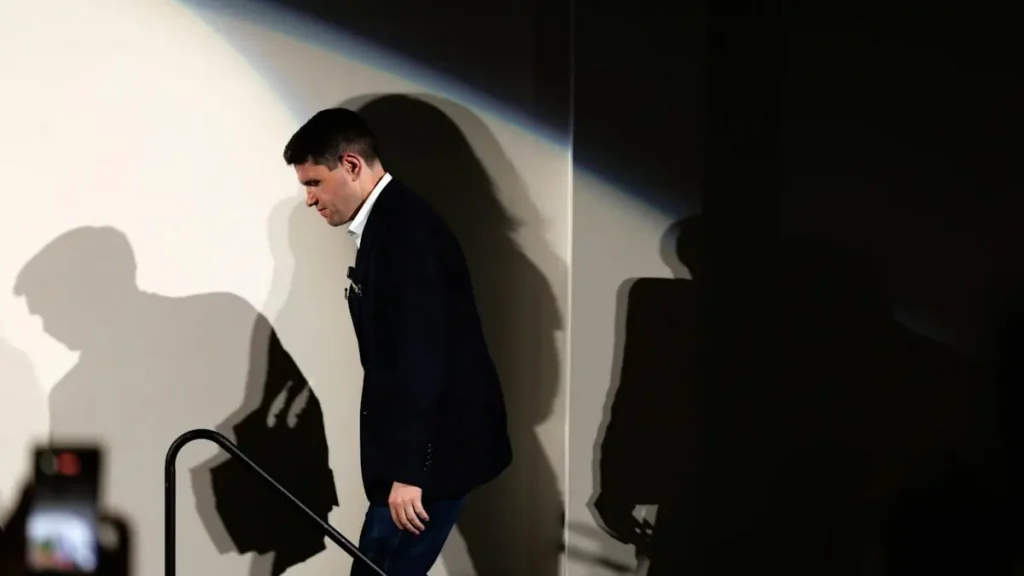 A man in a suit walks down a staircase, with his shadow cast on the wall behind him.