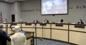The image shows a public meeting or hearing taking place in a conference room with a panel of officials seated at a long table, and a person standing at a podium addressing the panel.