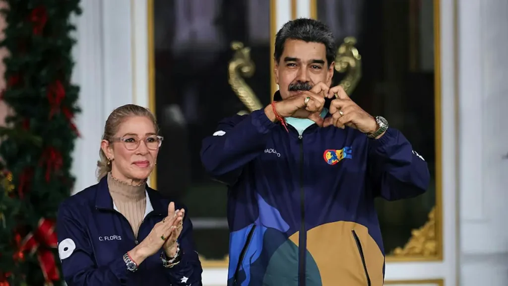 The image shows two individuals in matching jackets, one making a heart shape with their hands, in a setting decorated with Christmas elements.