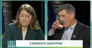 The image shows two individuals participating in a Vermont Public Debates 2026 event, with one person drinking from a glass and the other appearing to be listening or speaking.