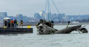 A helicopter is being recovered from the water by a crane, with a group of people observing the operation on a nearby platform.