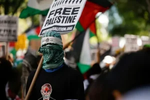 A protester wearing a keffiyeh and holding a sign that reads "FREE PALESTINE" and the website "ANSWERCOALITION.ORG" is seen at a demonstration.