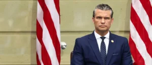A man in a suit and tie sits in front of two American flags.