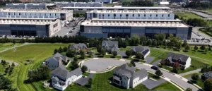 The image shows a suburban neighborhood with single-family homes surrounded by greenery, adjacent to a large industrial complex with multiple buildings and parking lots.