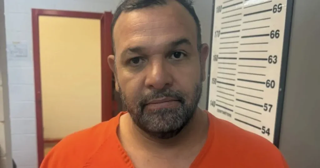 The image shows a person in an orange shirt standing in front of a height measurement chart, likely in a law enforcement or correctional facility setting.