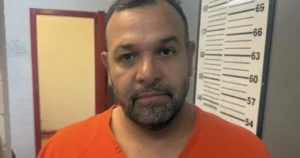 The image shows a person in an orange shirt standing in front of a height measurement chart, likely in a law enforcement or correctional facility setting.