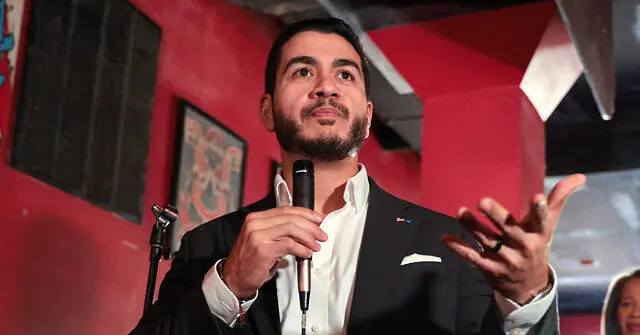 A man in a suit is speaking into a microphone, gesturing with his hands, in a setting with red walls and geometric shapes.