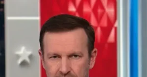 The image shows a close-up of a man in a news studio setting, with a red background featuring a pattern and a white star.