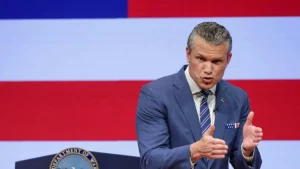 A man in a blue suit and tie is speaking in front of an American flag backdrop.