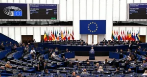 The image shows a formal meeting at the European Parliament, with a speaker addressing the assembly in front of the European Union flag and various national flags.