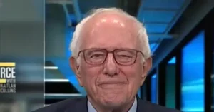 The image shows a man wearing glasses and a suit, appearing on a news program.
