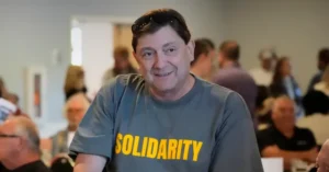 The photo shows a man wearing a shirt that says "SOLIDARITY" in a room with other people in the background.