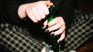 A person with dark nail polish is opening a green bottle, possibly a beer, while sitting on a checkered surface.
