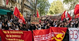 The photo depicts a large group of people marching in a protest, holding red flags and banners with slogans in French, advocating against imperialism and class war.