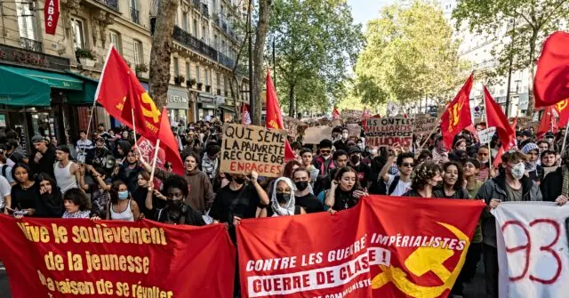 The photo depicts a large group of people marching in a protest, holding red flags and banners with slogans in French, advocating against imperialism and class war.