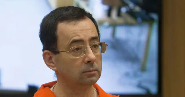 The image shows a person wearing an orange shirt, likely in a formal or official setting, with a blurred background that includes a monitor and other indistinct elements.