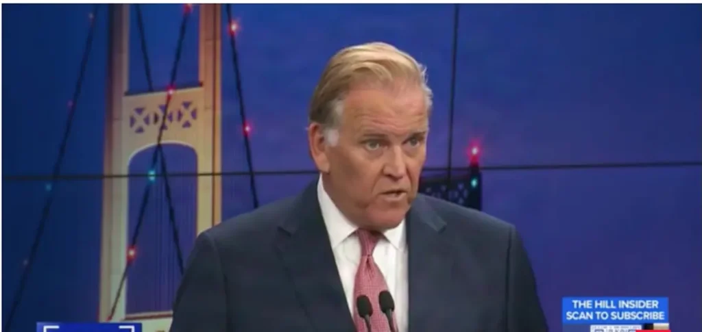 A man in a suit and tie is speaking on a news program, with a bridge and cityscape in the background.