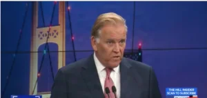 A man in a suit and tie is speaking on a news program, with a bridge and cityscape in the background.