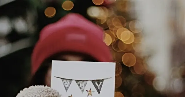 A festive scene featuring a person wearing a red hat and holding a card with a bunting design, set against a backdrop of blurred golden lights.