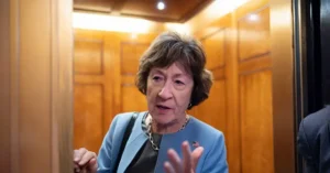 A woman in a blue blazer is seen inside an elevator, gesturing with her hands as she speaks.