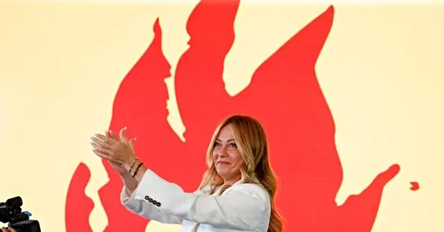 A woman in a white suit gestures with her hands in front of a backdrop featuring red flame-like shapes.